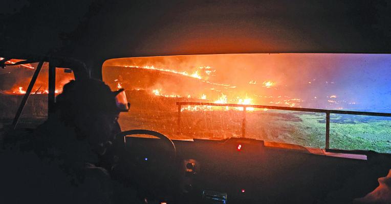A firefighter is silhouetted against the backdrop of a raging wildfire seen through the windshield of a fire truck. Recent fires have devastated a lot of the land in our area, leaving producers wondering what to do next. (Leader photo by Alisa Mullen)